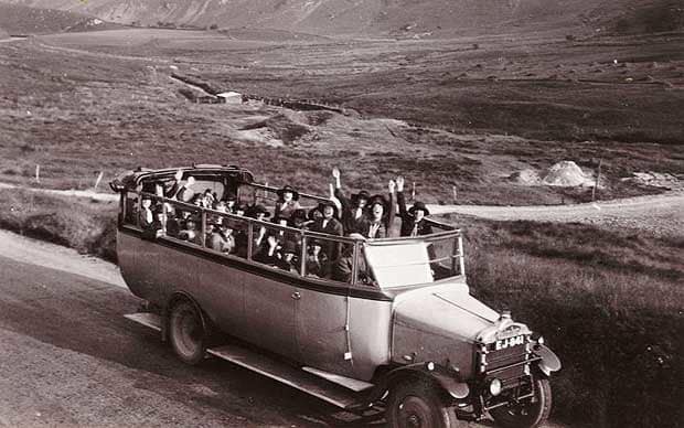 Black and white photo of an early, open top bus with people waving out the top