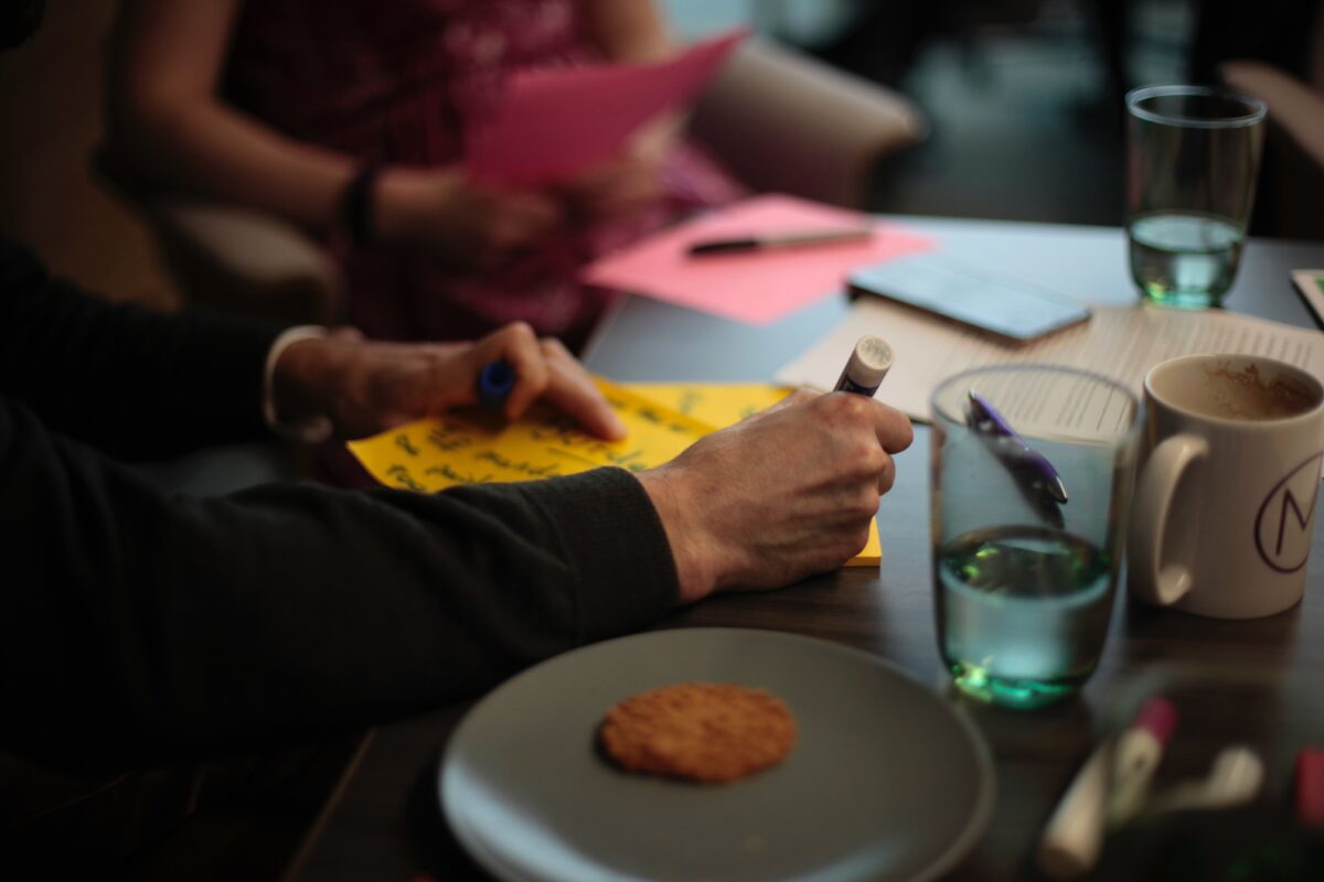 People working collaboratively using post-it notes. Their arms and torsos are visible. They are sat at a table with refreshments.