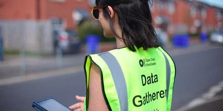 A photograph of a young white woman taken from behind, wearing sunglasses and a yellow high-vis jacket that says 'Open Data Manchester – Data Gatherer'