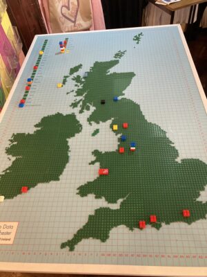 A photograph of Open Data Manchester's Happiness Index, which is a map of the Great Britain and the Republic of Ireland made of green lego board, on a blue background representing the sea, with extra slots in the top left representing the rest of the world. It has some coloured bricks, showing how happy certain people were and where they were happiest, dotted around the map.