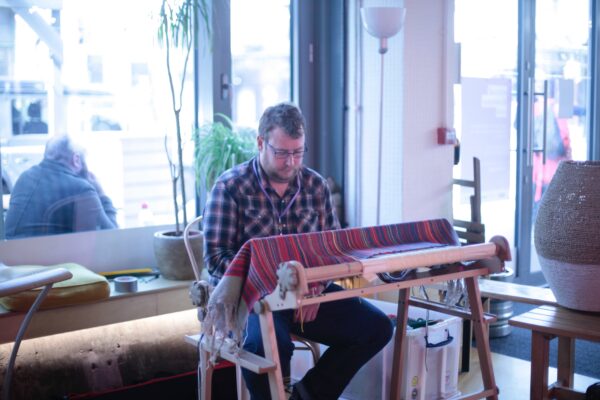 This is a picture of volunteer Tom sitting behind a wooden loom, weaving coloured thread representing people's show colour, into a piece of fabric