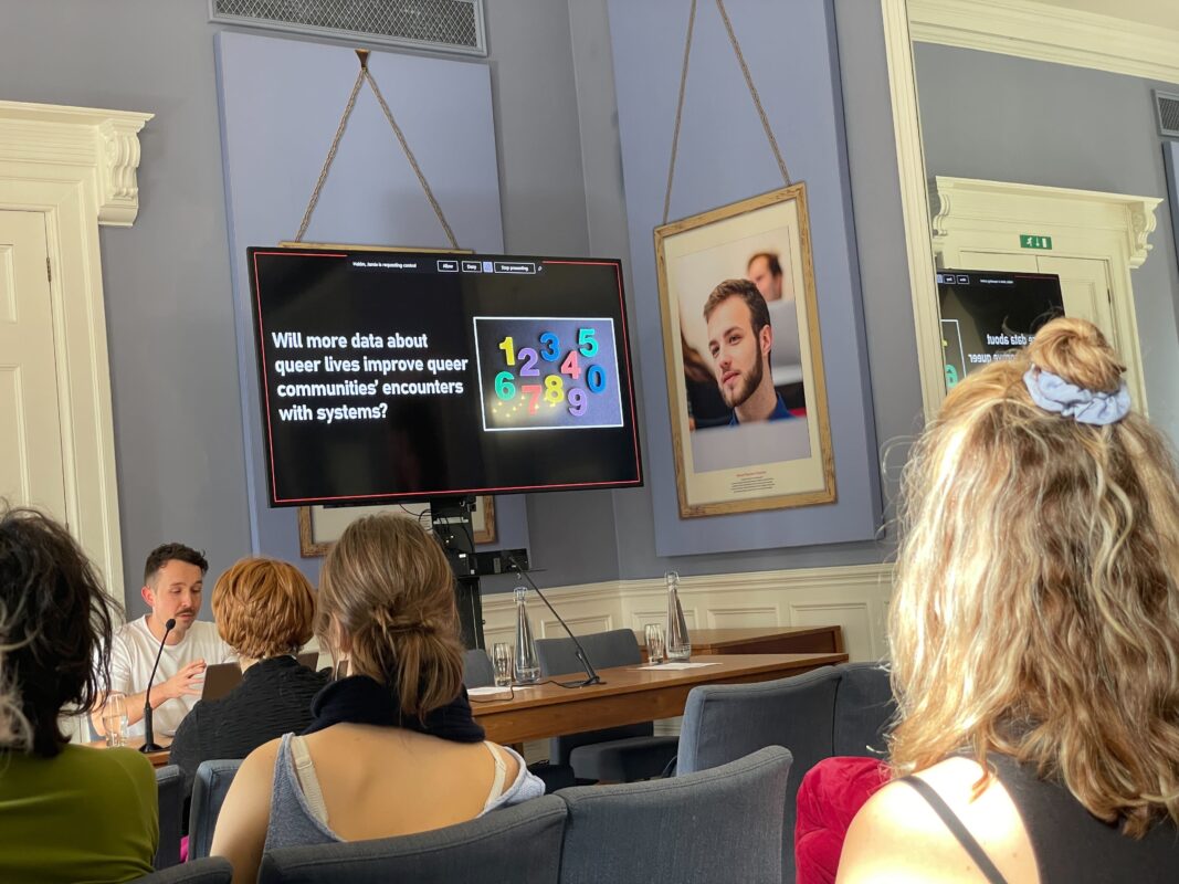 A white person delivering a talk to an audience, the slide displayed reads as follows: Will more data about queer lives improve queer communities' encounters with systems?