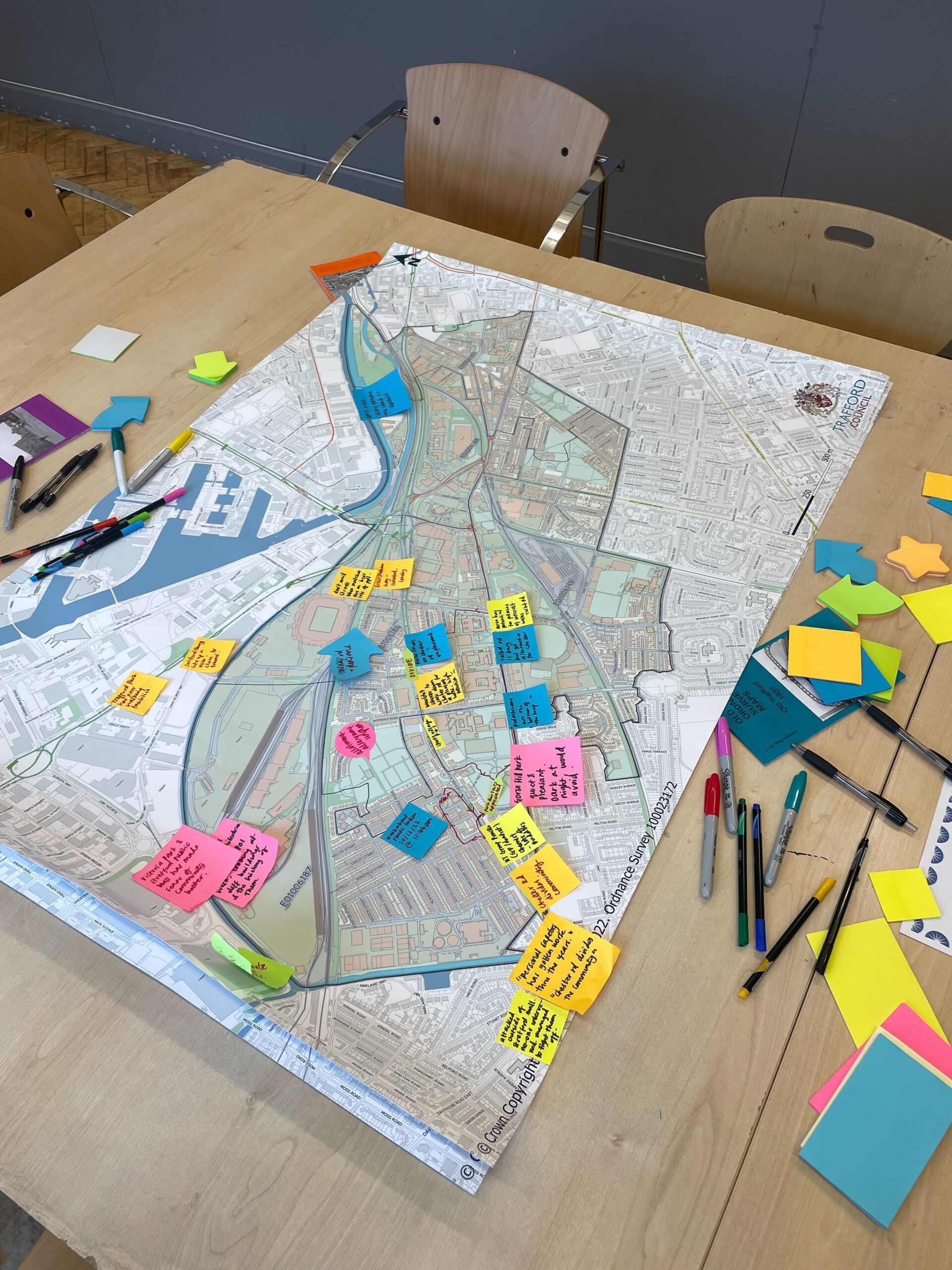 A map of Trafford on a wooden table, covered in post-it notes of different colours and surrounded by various pens.
