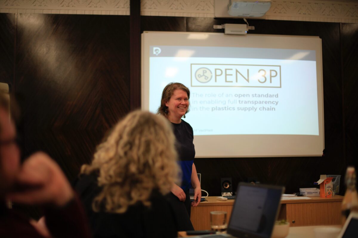 A white woman with medium length, wavy, brown hair, smiling ahead of delivering a talk. The slide displayed on the projector behind her reads Open 3P: The role of an open standard in enabling full transparency in the plastics supply chain.