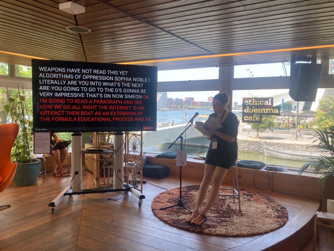 A white woman sitting on a high stool reading from a book, with her words being displayed on a large TV screen next to her