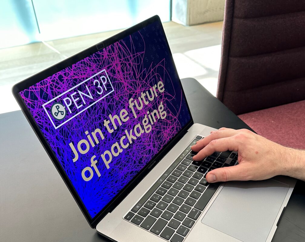 Laptop on desk with screen showing a purple background with swirly graduated patterns, with the Open 3P logo and Join the Future of Packaging in gold text. A hand is positioned on the right hand side of the image lightly touching the keyboard