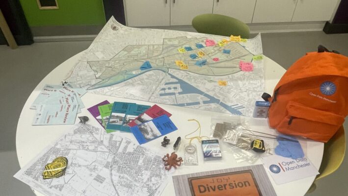 A round table in a classroom or office kitchen setting, covered with various items related to a mapping or urban planning project. The table contains: A large map with colored sticky notes attached Smaller printed maps and documents Colorful cards or badges An orange backpack with 'Open Data Manchester' logo Various small objects like figurines and measuring tools A prominent 'Diversion' sign image In the background, there's a kitchenette with white cabinets and a sink. On the left wall, there's a green board with 'ART' written on it, showing different categories. The scene suggests an interactive, collaborative project involving city planning or community mapping.