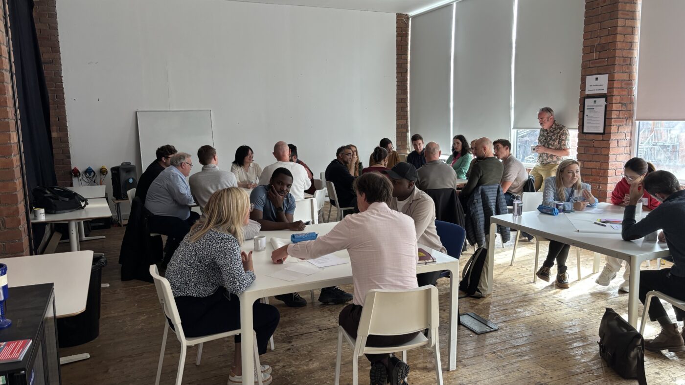 A room filled with data professionals sitting at white tables arranged in discussion groups. Participants are engaged in conversations during the September 2025 LCR Data Community workshop at DoES Liverpool, with exposed brick walls and industrial lighting visible in the background.