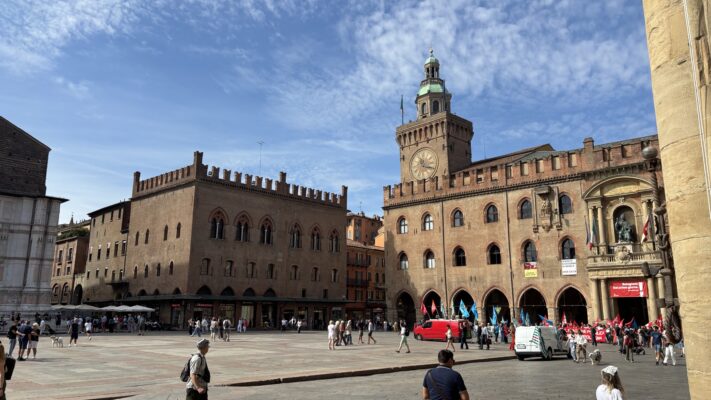Piazza Maggiore in Bologna, Italy, showing the medieval Palazzo d'Accursio (City Hall) with its distinctive clock tower topped with a green copper dome. The honey-colored brick building features Gothic arched windows and crenellations. The large plaza is filled with tourists and locals, with a small protest or demonstration visible with colorful flags. Adjacent medieval buildings line the square under a partly cloudy blue sky