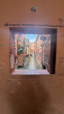 View through a small window in an ochre-colored wall covered with graffiti and visitor inscriptions, looking out onto Bologna's hidden canal system, the Canale delle Moline. The narrow waterway runs between tall pink and orange buildings with climbing ivy, reflecting the sky in its still waters. This is one of the few remaining visible sections of Bologna's historic underground canal network