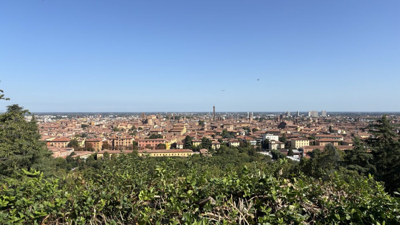 Panoramic view of Bologna, Italy, showing the historic city centre. The densely packed ochre, yellow, and pink buildings stretch to the horizon under a clear blue sky, with green foliage framing the foreground of this elevated viewpoint.